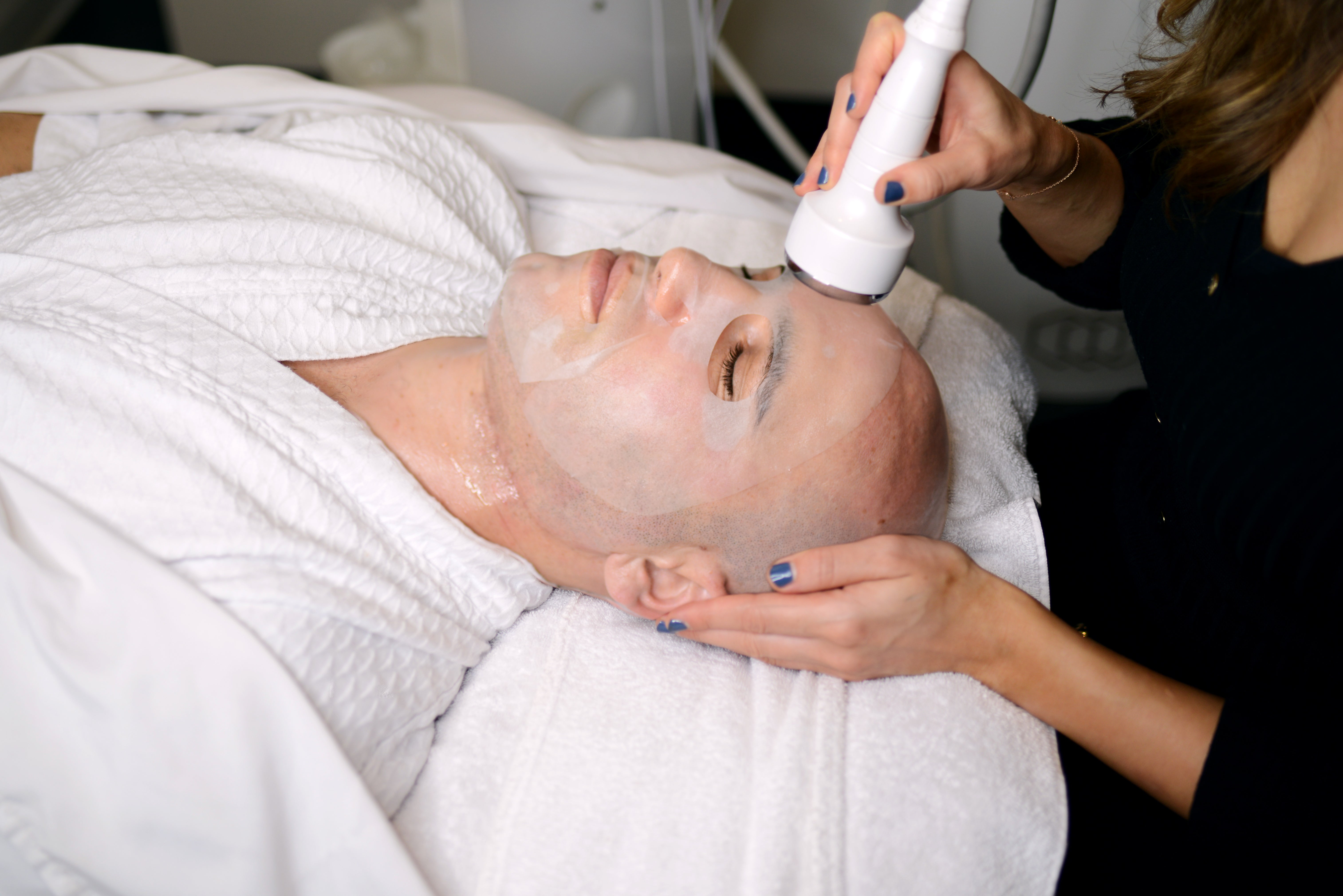 Man receiving facial treatment with handheld device while lying on white towel.